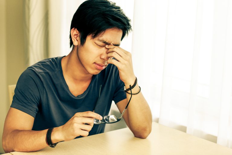 Tired asian man with eye pain holding glasses in hand